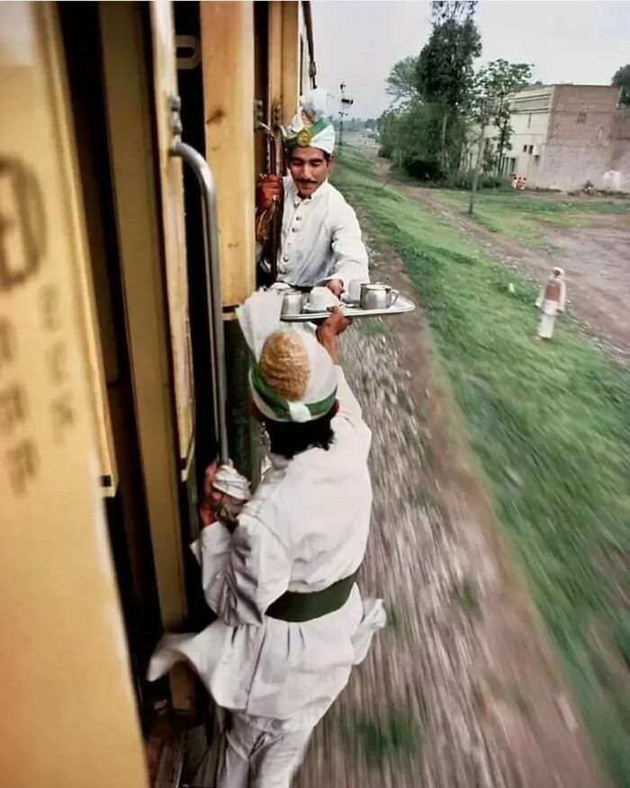 Two men passing breakfast tea from the kitchen car on a moving train, due to a door being locked between the train cars, on a railway from Peshawar to Rawalpindi, Pakistan, 1983.